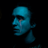 Na zdjęciu niebieska twarz artysty na tle czarnego tła. z lewego oka płynie czerwona łza.