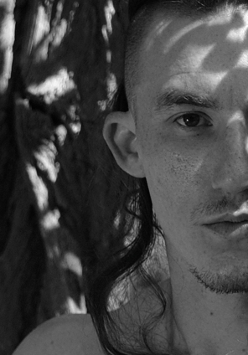 Czarno białe zdjęcie przedstawia twarz mężczyzny z lekkim zarostem i długimi włosami na tle drzewa.