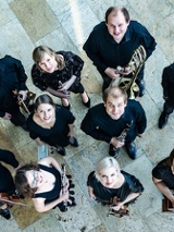 Na zdjęciu wykonawcy koncertu z instrumentami stoją obok siebie i zadzierają głowy w górę, w stronę obiektywu aparatu.