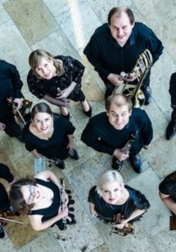 Na zdjęciu wykonawcy koncertu z instrumentami stoją obok siebie i zadzierają głowy w górę, w stronę obiektywu aparatu.
