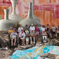 Zdjęcie przedstawia grupę osób z niepełnosprawnościami znajdujących się na pustkowiu. W tle widoczne są olbrzymie wazy, widoczne są także ślady farby.