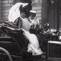 kadr z czarno-białego filmu "Kobiety za kierownicą"; na siedzeniu samochodu prowadzonego przez szofera siedzi kobieta w białej sukni trzymająca w ręku parasol