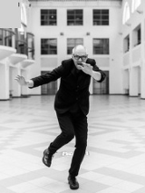 Czarno-biała fotografia. W centralnym punkcie Mariusz Lubomski w ruchu, w czarnym garniturze i okularach na nosie. Pozuje na tle dużego, jasnego korytarza, który z każdej strony jest otoczony oknami i portalami.