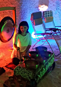 Zdjęcie przedstawia kobietę grającą na misach; za jej plecami widoczny jest gong oraz leżaki.