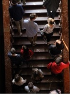 Grupa ludzi ukazana z perspektywy lotu ptaka wchodzi po schodach. Przed nimi okno; świetlają ich promienie zachodzącego słońca.