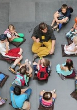 Grupa dzieci oraz osoba dorosła - przewodnik - siedzą na poduszkach rozłożonych na szarej kamiennej posadzce. Zdjęcie wykonane z perspektywy lotu ptaka.