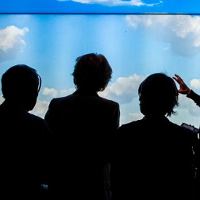 Ludzie stojący przodem do ekranu wyświetlającego chmury. Przewodniczka wskazuje rękoma i prezentująca wystawę.