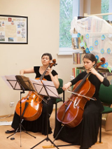 Na zdjęciu dwie siedzące kobiety, grające na wiolonczelach.