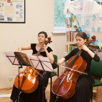 Na zdjęciu dwie siedzące kobiety, grające na wiolonczelach.