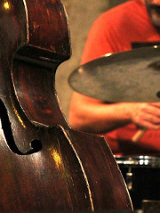Zdjęcie na którym widać część kontrabasu. w tle muzyk przy perkusji.