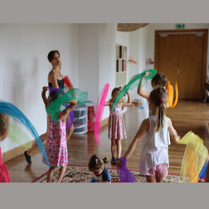 Zdjęcie przedstawia kobietę i grupkę małych dzieci trzymających kolorowe kawałki zwiewnej tkaniny.