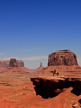 Kadr pochodzi z westernu Johna Forna "Nosiła żółtą wstążkę" i przedstawia panoramiczny widok na ikoniczną górę Oljato w Monument Valley w Colorado. Niebieskie niebo, czerwono-brunatne skały i piasek, na środku kadru widać kowboja na koniu, stojącego na małym wzniesieniu.