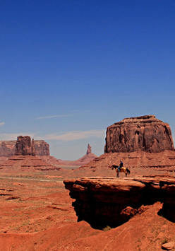 Kadr pochodzi z westernu Johna Forna "Nosiła żółtą wstążkę" i przedstawia panoramiczny widok na ikoniczną górę Oljato w Monument Valley w Colorado. Niebieskie niebo, czerwono-brunatne skały i piasek, na środku kadru widać kowboja na koniu, stojącego na małym wzniesieniu.
