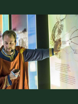 Przewodnik przebrany w średniowieczny strój stoi przy interaktywnej tablicy, na której widoczne są nazwy wielkopolskich miast, m.in. Poznań, Gniezno.