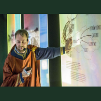 Przewodnik przebrany w średniowieczny strój stoi przy interaktywnej tablicy, na której widoczne są nazwy wielkopolskich miast, m.in. Poznań, Gniezno.