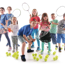 Dzieci z rakietami tenisowymi otoczone na podłodze piłkami do niego.