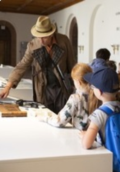 Zdjęcie detektywa w kapeluszu, a obok niego grupka dzieci - uczestników warsztatów.