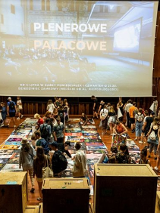 Drewniania sala z rozłożonymi plakatami na ziemi, a wśród nich ludzie wybierający je.