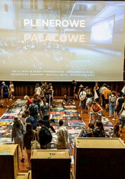 Drewniania sala z rozłożonymi plakatami na ziemi, a wśród nich ludzie wybierający je.