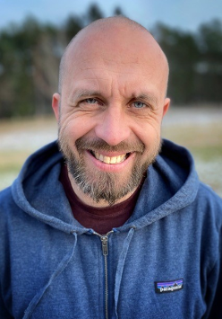 Zdjęcie Roberta Maciąga. Stoi uśmiechnięty. Głowa łysa, niebieska bluza z opuszczonym na ramiona kapturem.