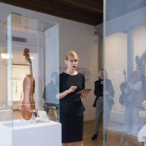Zdjęcie Pani oprowadzającej po wystawie w Muzeum Instrumentów Muzycznych. Widać szklane gabloty z instrumentami umieszczonymi wewnątrz.
