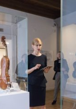 Zdjęcie Pani oprowadzającej po wystawie w Muzeum Instrumentów Muzycznych. Widać szklane gabloty z instrumentami umieszczonymi wewnątrz.