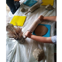 Na zdjęciu dziecięce ręce rozwałkowują glinę wałkiem na stole. Obok stoi puszka z patyczkami.