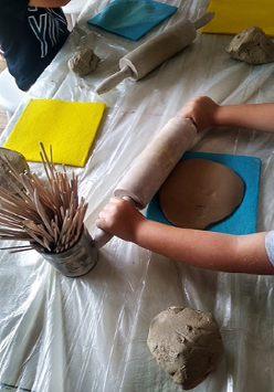 Na zdjęciu dziecięce ręce rozwałkowują glinę wałkiem na stole. Obok stoi puszka z patyczkami.