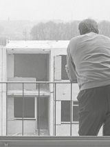 fotografia przedstawia, mężczyznę odwróconego plecami, opierającego się o barierkę balkonu, w tle widać niedokończony wielopiętrowego budynek