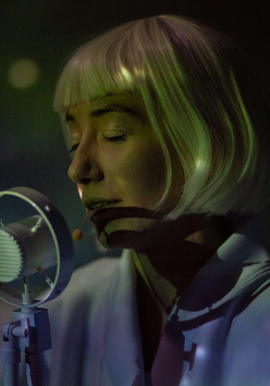 na zdjęciu twarz kobiety o jasnych włosach, z grzywką, przed nią mikrofon. Kobieta ma zamknięte oczy.