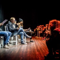 Na zdjęciu fragment przedstawienia. Na scenie, twarzami do widzów, na krzesłach siedzą aktorzy, czytają.