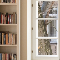 Na fotografii wnętrze Filii Sztuki Biblioteki Raczyńskich. Widać białe regały książkowe i duże okno, za którym widać fragment pnia i gałęzi platana.