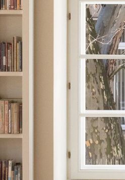 Na fotografii wnętrze Filii Sztuki Biblioteki Raczyńskich. Widać białe regały książkowe i duże okno, za którym widać fragment pnia i gałęzi platana.