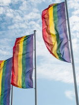 Zdjęcie 3 flag LGBT na masztach na tle niebieskiego nieba z białymi chmurkami.