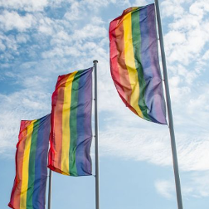 Zdjęcie 3 flag LGBT na masztach na tle niebieskiego nieba z białymi chmurkami.