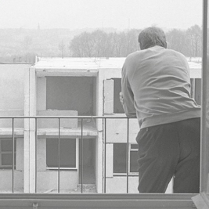 fotografia przedstawia, mężczyznę odwróconego plecami, opierającego się o barierkę balkonu, w tle widać niedokończony wielopiętrowego budynek