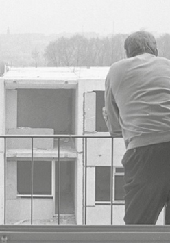 fotografia przedstawia, mężczyznę odwróconego plecami, opierającego się o barierkę balkonu, w tle widać niedokończony wielopiętrowego budynek