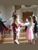 Na zdjęciu grupa dzieci wraz z prowadzącą zajęcia tańczy trzymając się za ręce w kole.