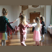 Na zdjęciu grupa dzieci wraz z prowadzącą zajęcia tańczy trzymając się za ręce w kole.
