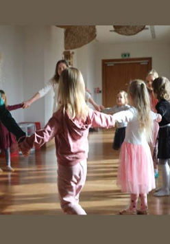 Na zdjęciu grupa dzieci wraz z prowadzącą zajęcia tańczy trzymając się za ręce w kole.