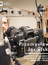 W centralnej części zdjęcia widać maszynę fotografującą. W tle stoi mężczyzna w okularach i wpatruje się w obiektyw. W prawym dolnym rogu widnieje tytuł wystawy i nazwisko autora.