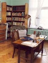 Wnętrze mieszkania-pracowni poetki. Na pierwszym planie stół zastawiony dokumentami, w tle regał z książkami.