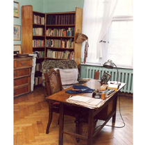 Wnętrze mieszkania-pracowni poetki. Na pierwszym planie stół zastawiony dokumentami, w tle regał z książkami.