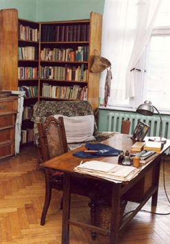 Wnętrze mieszkania-pracowni poetki. Na pierwszym planie stół zastawiony dokumentami, w tle regał z książkami.
