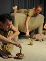 Zdjęcie aktorów w trakcie spektaklu. Trzech aktorów ubranych na beżowo z młynkowymi instrumentami. Beżowy słonik z różowymi uszami.