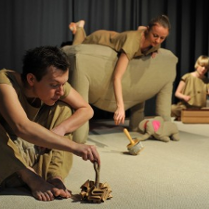 Zdjęcie aktorów w trakcie spektaklu. Trzech aktorów ubranych na beżowo z młynkowymi instrumentami. Beżowy słonik z różowymi uszami.