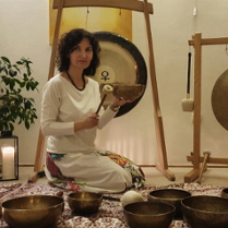 Kobieta siedzi na dywanie, w dłoni trzyma misę i gong. Na dywanie stoją złote misy, w tle duży gong.