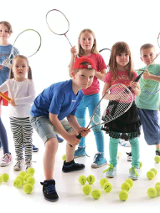 Dzieci z rakietami tenisowymi otoczone na podłodze piłkami do niego.