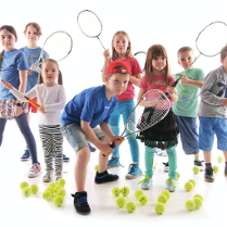 Dzieci z rakietami tenisowymi otoczone na podłodze piłkami do niego.