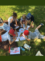 Grupa dzieci siedzi na kocu na trawie i rysuje kolorowymi kredkami na kartkach.